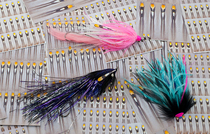 Close-up of three Aqua Flies Un-Real Jungle Cock fishing lures in pink, purple, and teal with feather and synthetic materials, designed for musky fishing tackle.