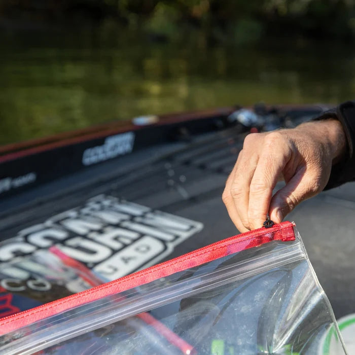 Hand holding the zipper of a transparent red and clear Bass Mafia Money Bag on a fishing boat with fishing rods and tackle in the background.
