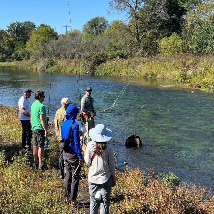 Group of anglers standing on a riverbank learning fly fishing techniques with fishing rods in hand during Bob Mitchell's Fly Fishing College near clear flowing water and green trees.