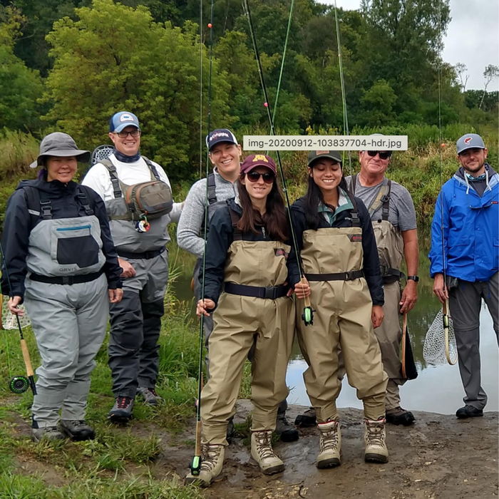 Group of six diverse anglers standing by a riverbank wearing fishing waders and gear, holding rods and nets, participating in Bob Mitchell's Fly Fishing College outdoors.