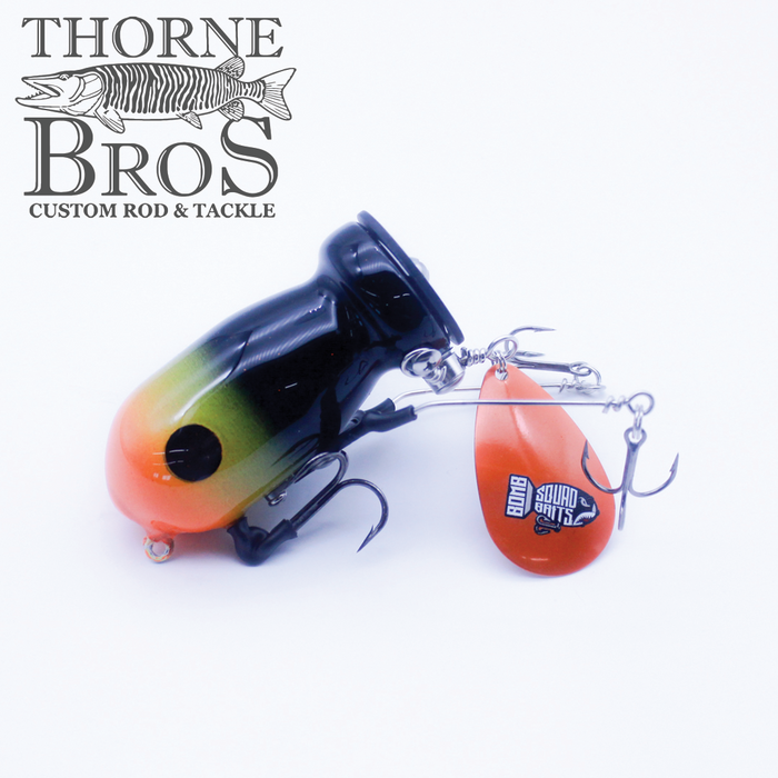 Close-up of a Bomb Squad C-4 Flap Tail fishing lure featuring a black, green, and orange body with a large treble hook and an orange spinner blade designed for musky fishing tackle.