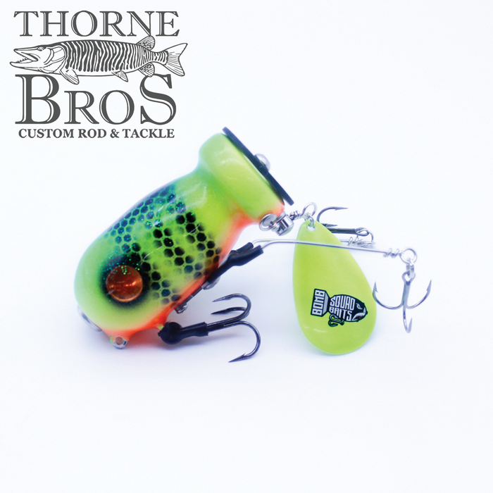 Brightly colored fishing lure with green, yellow, and orange hues, featuring black scale patterns, two treble hooks, and a green spinner blade labeled Bomb Squad C-4 Flap Tail.