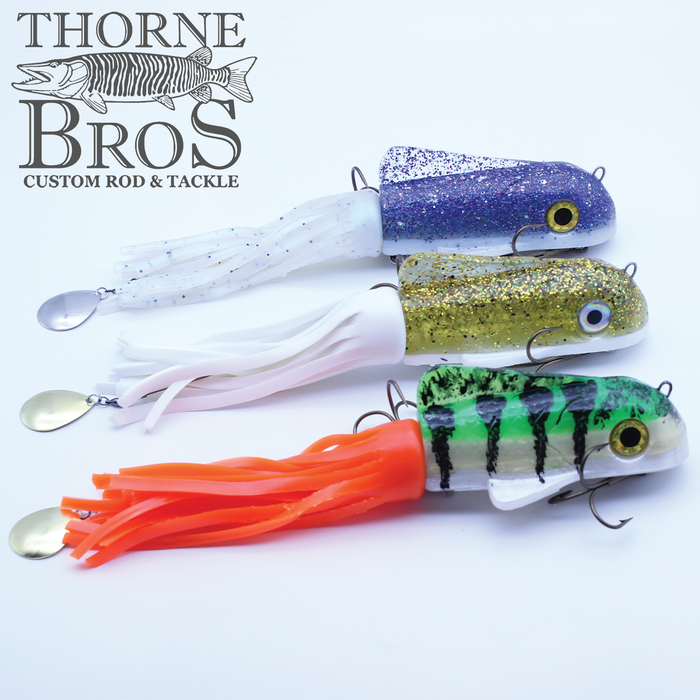 Three Boss Shad Large Hybrid fishing lures in purple glitter with white tail, gold glitter with white tail, and green with black stripes and orange tail, each featuring hooks and metal spinner blades, displayed on a white background with Thorne Bros Custom Rod & Tackle logo.