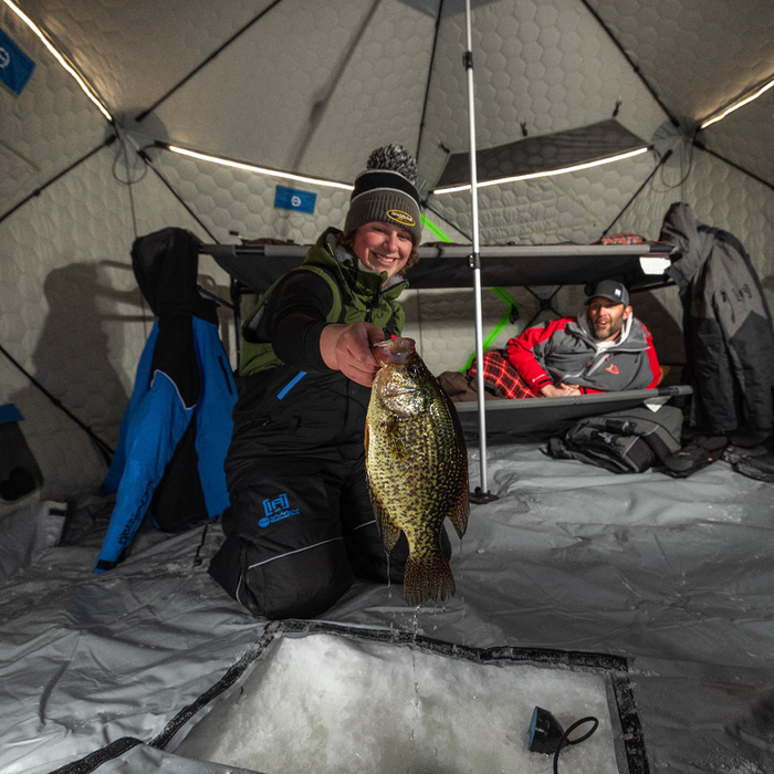 Two people inside an insulated ice fishing shelter illuminated by Clam Aurora LED Light Kits, with one person holding a freshly caught fish over an open ice fishing hole.