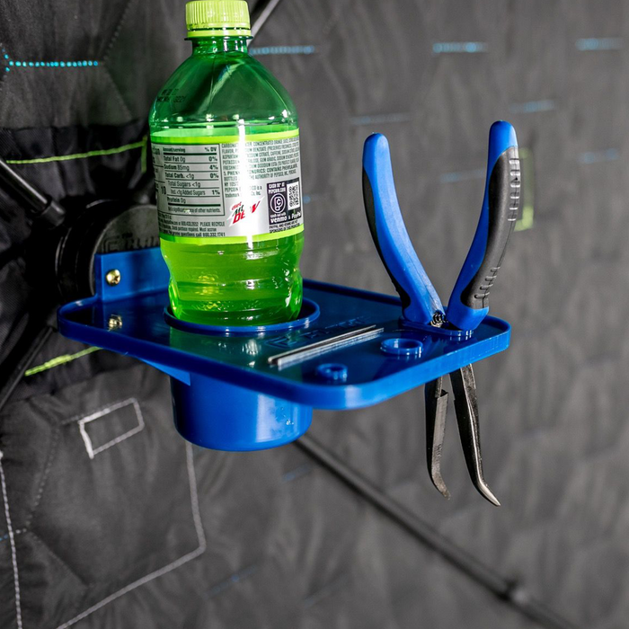 Clam ClamLock Hub Shelf in blue securely holds a green beverage bottle, pliers with blue handles, and small fishing accessories on a quilted fabric background.