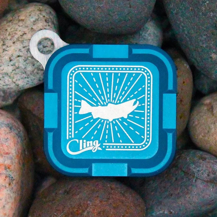 Square blue Cling Mag Grab Plus magnetic tackle box with a fish design on the lid, placed on smooth river rocks, designed for organizing fishing tackle.