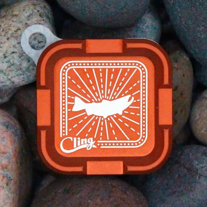 Orange square compact container labeled Cling Mag Grab Plus with a white fish graphic, placed on smooth grey and beige fishing rocks.