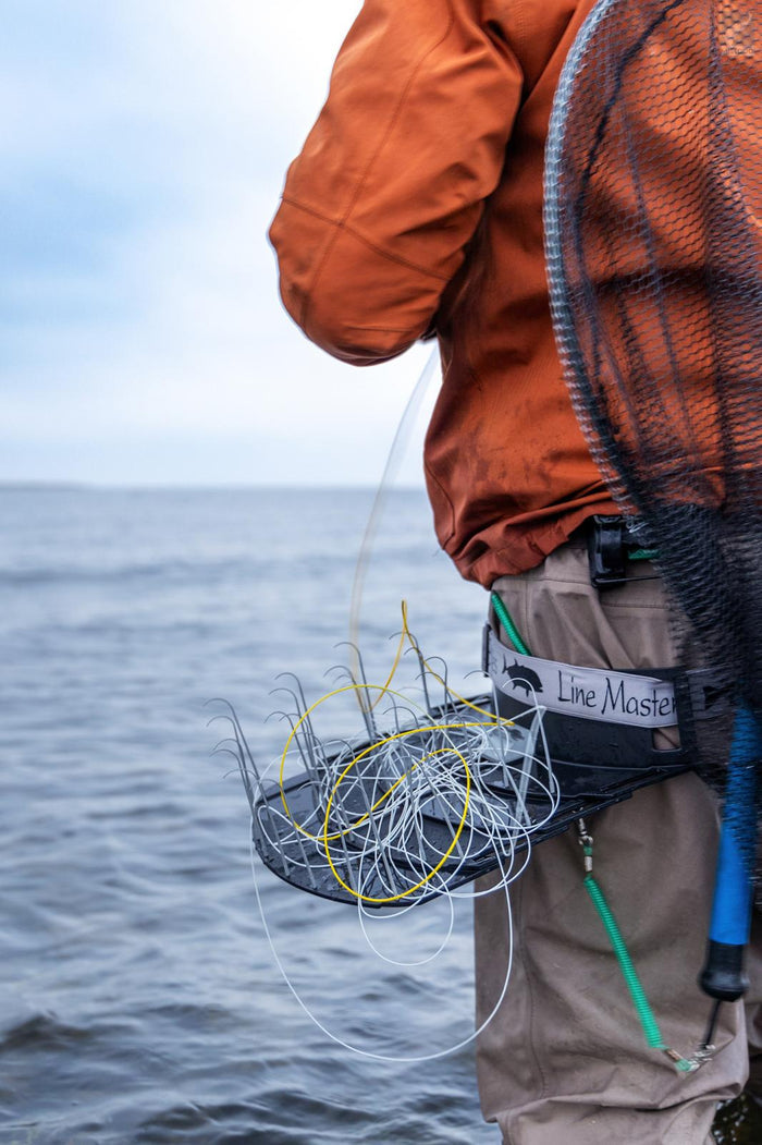 Person wearing an orange jacket and beige pants standing by water, equipped with an ESE Line Master Stripping Basket holding fishing lines, alongside a fishing net attached to the waist.