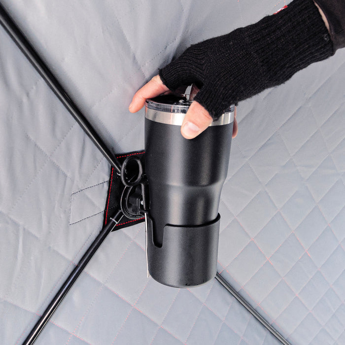 Hand wearing a black fingerless glove placing a black insulated tumbler into a built-in holder of the Eskimo E-Hub Cup Holder attached to a gray insulated surface, designed for secure beverage placement during outdoor fishing activities.