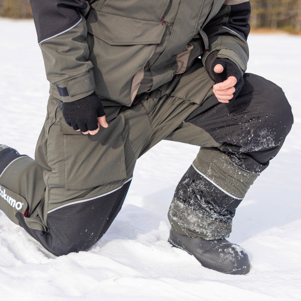 Man wearing Eskimo Expedition Bib - Men's, designed for cold weather fishing, kneeling in snow, featuring durable, insulated fabric with reinforced knees and waterproof boots.