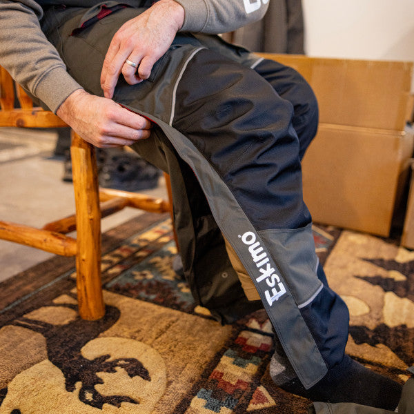 Close-up of a person sitting on a wooden chair wearing black and gray Eskimo Expedition Bib - Men's fishing bib pants with white logo, indoors on a patterned rug.