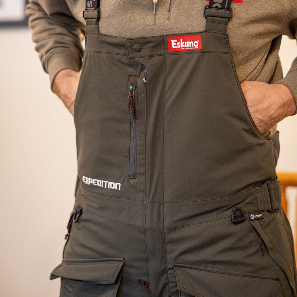 Close-up of a person wearing Eskimo Expedition Bib - Men's in dark olive, featuring front zipper pocket, multiple cargo pockets, adjustable shoulder straps, and durable outdoor fabric for fishing and ice expeditions.
