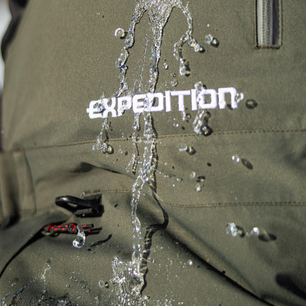 Water droplets running off close-up of olive green Eskimo Expedition Bib - Men's showing waterproof fabric and embroidered "EXPEDITION" text on chest.