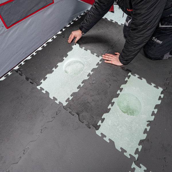 Person kneeling inside an ice fishing shelter, installing dark interlocking Eskimo Floor Tile Kits around two rectangular cutouts in the ice for fishing holes.