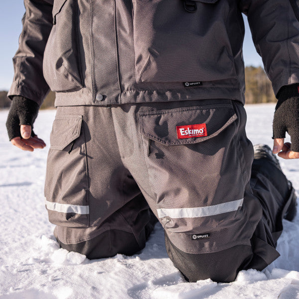 Eskimo Roughneck Bib - Men's in dark gray, insulated and waterproof with multiple cargo pockets and reflective strips, worn kneeling on snow for ice fishing.