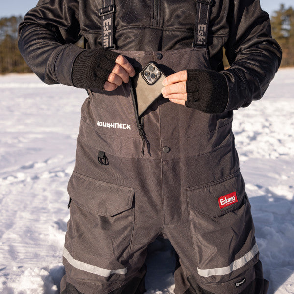 Man wearing Eskimo Roughneck Bib - Men's in dark gray with reinforced knees and multiple pockets, using a front zipper pocket outdoors in snowy landscape.