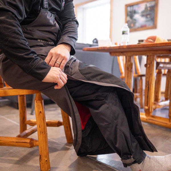 Man seated on wooden chair indoors wearing black Eskimo Roughneck Bib - Men's, shown with zippered side opening and adjustable ankle cuffs.
