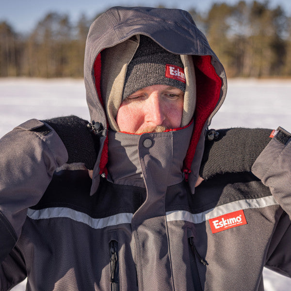 Man outdoors wearing a gray and black Eskimo Roughneck Jacket - Men's with a hood and red lining, adjusting the hood on a snowy landscape, dressed for cold weather fishing.