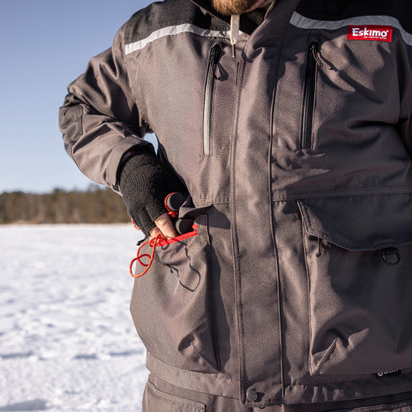 Man wearing a gray Eskimo Roughneck Jacket - Men's with multiple zippered and flap pockets, adjusting gear while standing on a snowy outdoor ice fishing area.