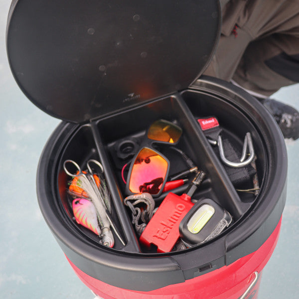 Eskimo Seatstash Bucket Lid shown open on a red bucket containing fishing tackle including hooks, sunglasses, a lure, scissors, and a red Eskimo branded item for organized fishing gear storage.