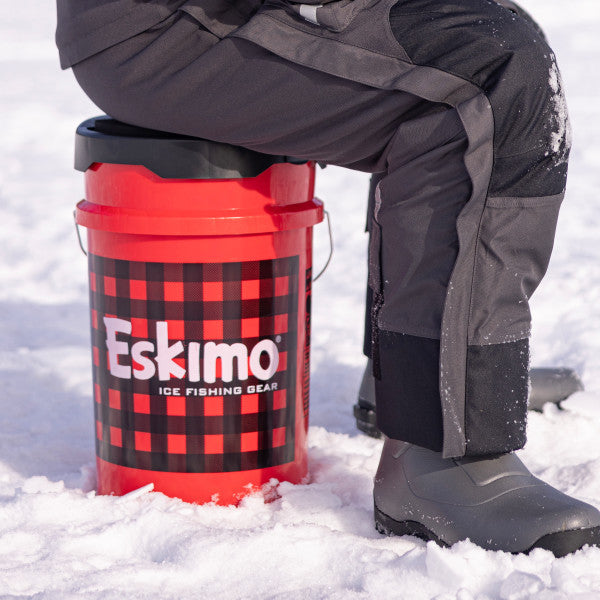 Person wearing black snow pants and boots sitting on a red and black plaid bucket with Eskimo logo, featuring the Eskimo Seatstash Bucket Lid on snowy ice fishing terrain.