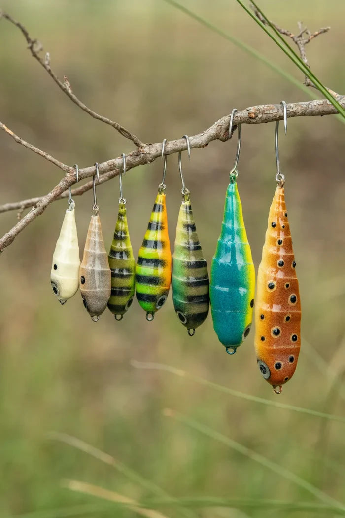 Seven colorful Fish Chase Flies Spiral Spook fishing lures in various patterns and sizes hanging from a branch, designed for fishing tackle enthusiasts.