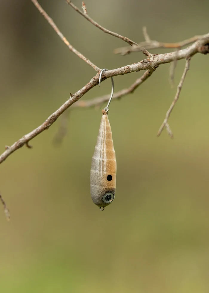 Fish Chase Flies Spiral Spook fishing lure in tan and brown hues with black spot and lifelike eye, hanging from a tree branch against a blurred natural background.
