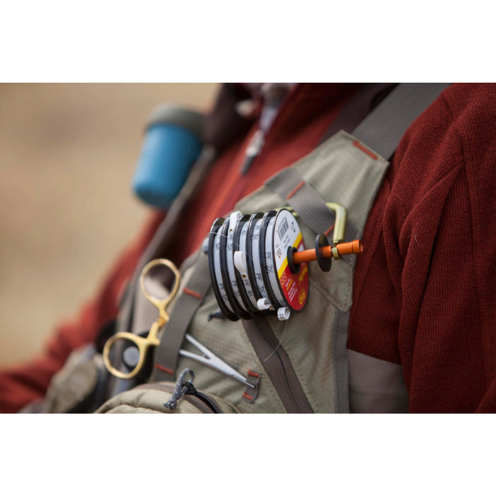 Fishpond Headgate Tippet Holder attached to a fishing vest, holding multiple spools of fishing tippet for fly fishing, with a pair of fishing forceps clipped nearby.