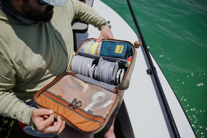 Man on boat holding an open Fishpond Honcho Reel Case filled with organized fishing lines, leaders, and tackle gear, next to a fishing rod over green water.