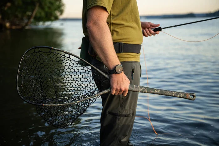 Angler standing in shallow water holding a Fishpond Nomad Middle Fork Net with a camo-patterned handle and rubber mesh netting, preparing for fly fishing.