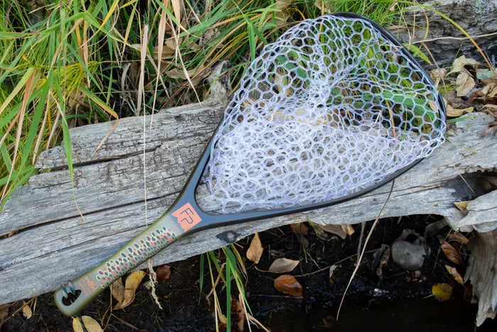 Fishpond Nomad Yampa Hand Net with a clear rubber mesh net and patterned handle resting on weathered wood near grass and water, designed for fishing tackle use.