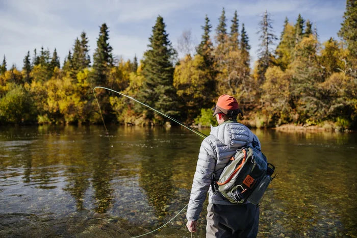 Angler wearing a gray jacket and orange cap fishing in a clear river with evergreen trees in the background, carrying a Fishpond Stormshadow Sling Pack on their back.