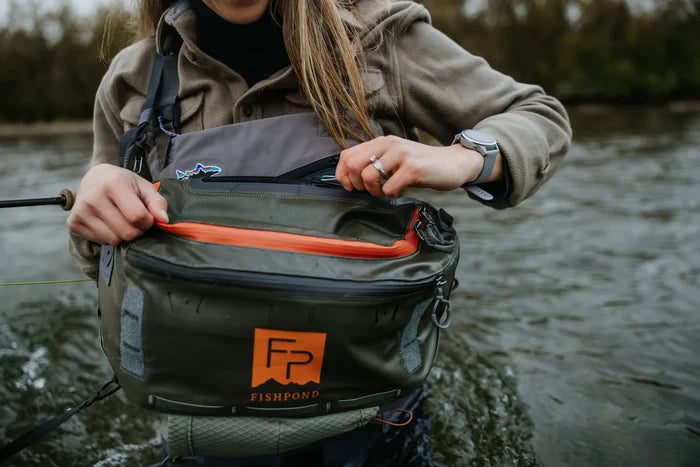 A person wearing a grey jacket and watch opens the front zippered pocket of an olive green Fishpond Stormshadow Sling Pack with orange accents while standing in a river with fishing gear.