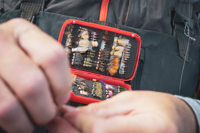 Fulling Mill Fly Patch Box open to display organized colorful fishing flies inside, held by an angler wearing outdoor fishing gear preparing a fly fishing rig.