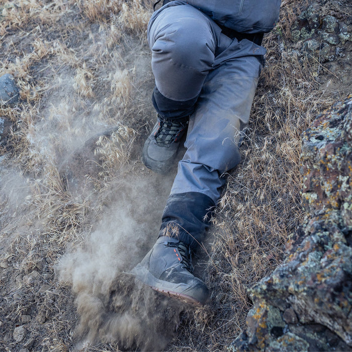 Close-up of a person wearing Grundens Bedrock Wading Boot-Rubber Sole climbing a rocky, dry terrain with dust rising from the boot's rugged sole.