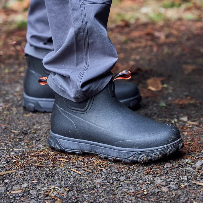 Grundens Deviation Sherpa Ankle Boot in black, shown on a person standing outdoors on a dirt and leaf-covered ground, featuring a durable, waterproof design with textured sole for traction.