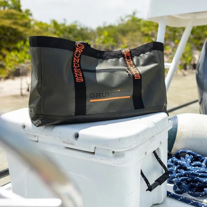 Grundens Gear Hauler Tote Bag 50L in dark green with black handles and orange logo detailing, placed on a boat cooler with marine equipment in the background.