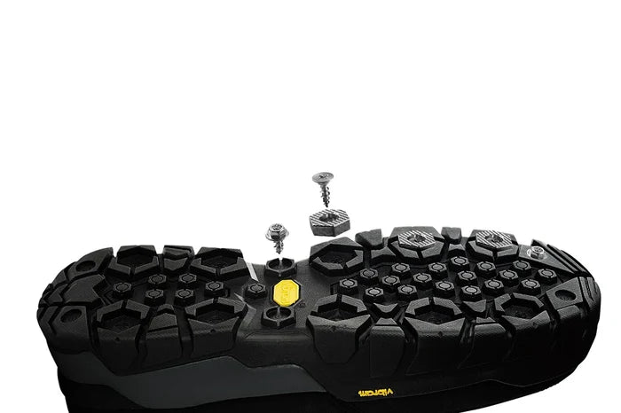 Black rugged sole of a footwear with textured grip pattern featuring silver metal cleats and studs detached above the sole, designed for added traction and durability in outdoor conditions, displayed with Grundens Hexagrip Cleat and Stud Pack.