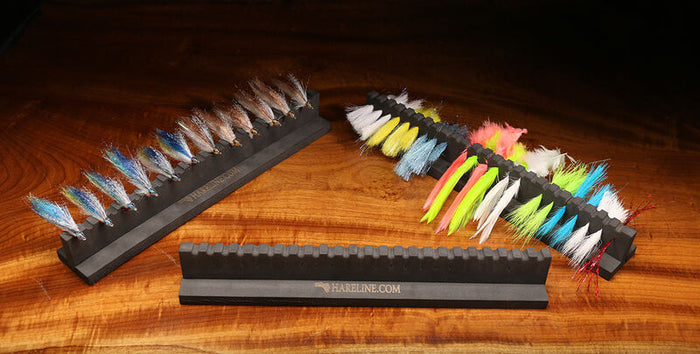 Hareline Chicone's Material & Fly Prep Station displaying multiple colorful fishing flies organized in a black foam holder on a wooden surface, used for fly fishing tackle preparation.