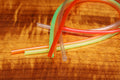 Several colorful translucent tubes including orange, green, and clear are arranged on a wooden surface, illustrating Hareline Hook Holder Tubing used in fishing tackle setups.