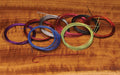 Various coils of colored wire neatly arranged on a wooden surface, including purple, blue, gray, red, green, black, and brown, representing the Hareline Senyo Intruder Wire used for fishing tackle customization.