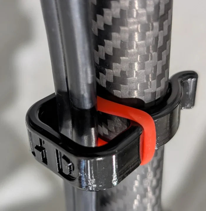 Close-up of a black HD Innovations HD Pole Clips (3-Pack) securing two fishing rods against a carbon fiber fishing rod, featuring a red flexible band for added stability.