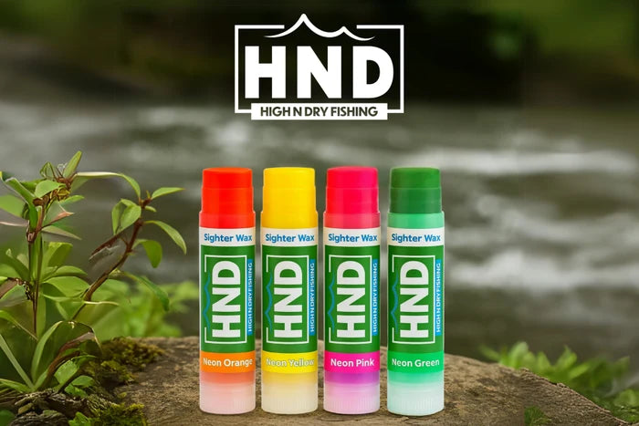 Four tubes of High and Dry Sighter Wax in neon orange, neon yellow, neon pink, and neon green colors standing upright on a mossy rock near a flowing river, with the HND High N Dry Fishing logo above.