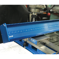 Ketch X Bump Board precision measuring tool in blue, set up in a workshop with a machining vise and drill press, used for accurately measuring fishing tackle components.