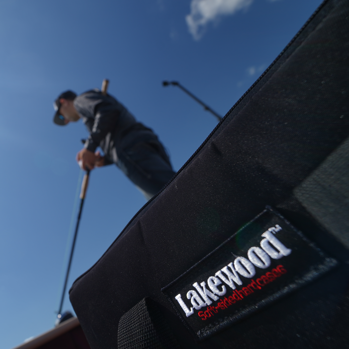 Close-up of a black padded fishing tackle bag labeled Lakewood with a blurred angler holding a fishing rod in the background, showcasing the Lakewood Musky Box - Shallow Invader fishing tackle storage solution.