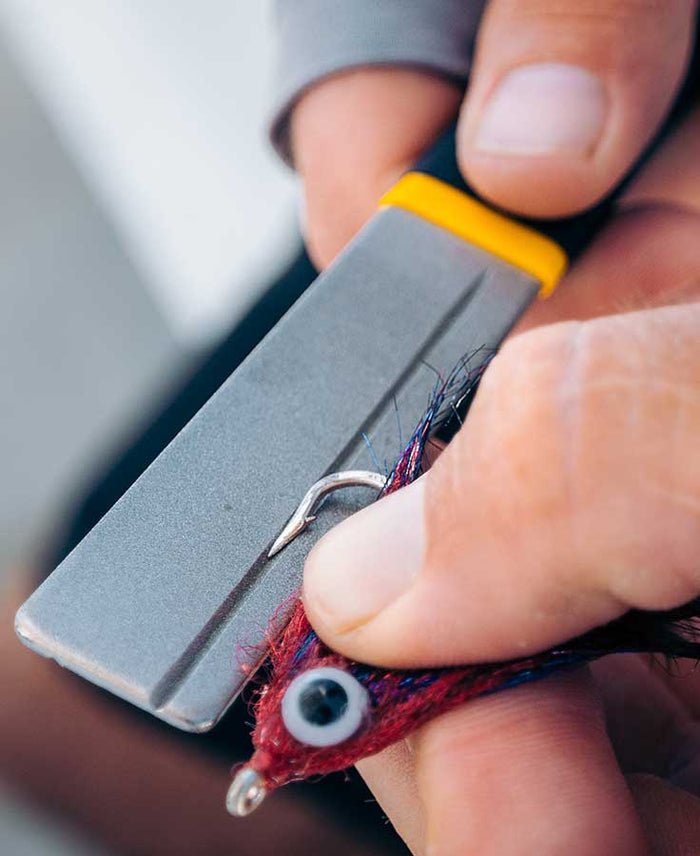 Close-up of hands using a Loon Apex Hook Hone XL with a yellow and black handle to sharpen the hook on a red fishing lure, emphasizing precision in fishing tackle maintenance.
