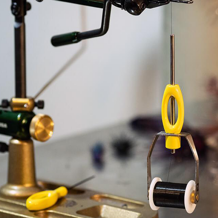 Close-up of a yellow Loon Ergo Bobbin fishing line tensioner hanging from a fishing rod setup, with a spool of black fishing line and a yellow-handled tool nearby.