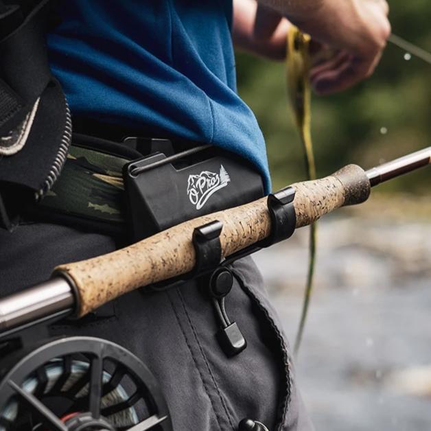 O'Pros Belt Clip Rod Holder attached to a fisherman's belt securing a fly fishing rod with a cork handle, showing hands casting a line over a river background.