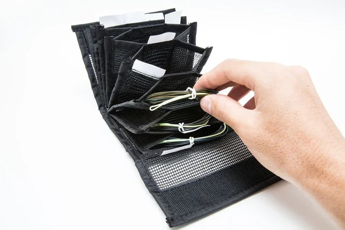 A hand holding a green fishing line segment inside an open black OPST Sink Tip Wallet with multiple mesh and fabric compartments for organized fishing tackle storage.