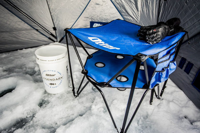 Otter Deluxe Double Decker Table - #201622 in blue with dual surfaces, cup holders, and side storage pocket, set up on ice inside an ice fishing shelter with a branded cup and gloves nearby.